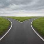 A two-lane road splitting into two directions surrounded by green fields under a cloudy sky.